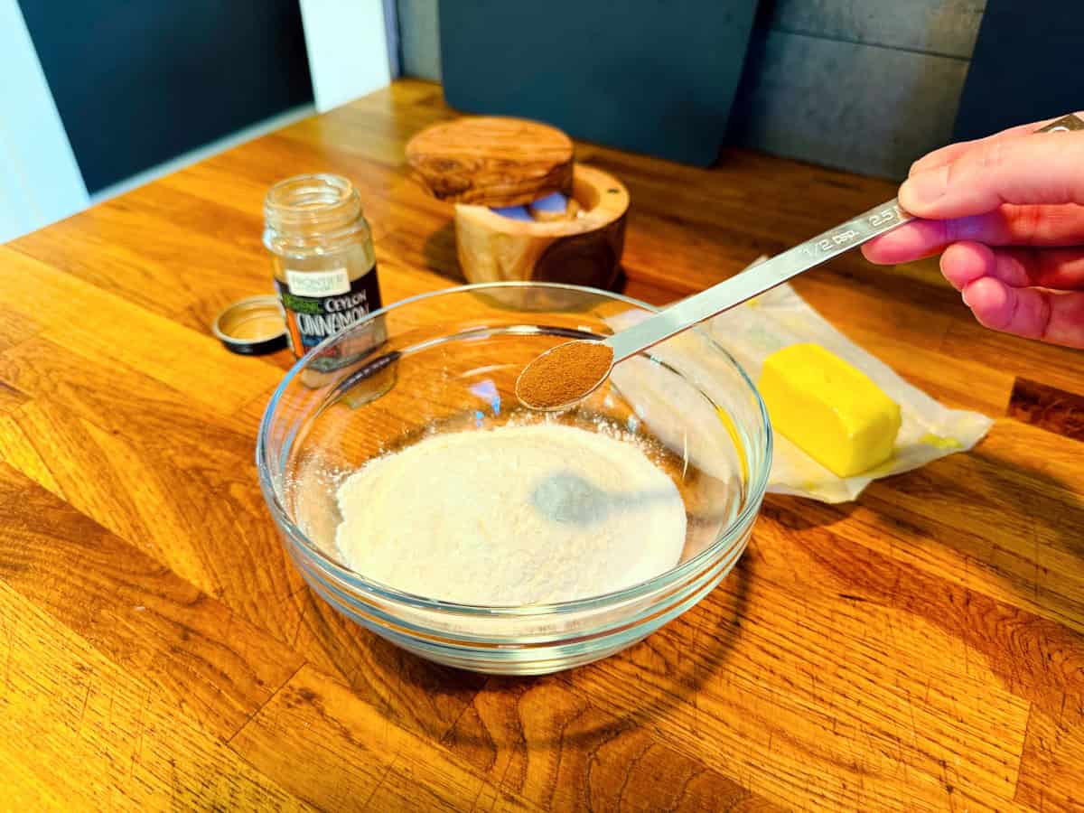 Cinnamon in a steel measuring spoon being held over a small bowl of flour and sugar.