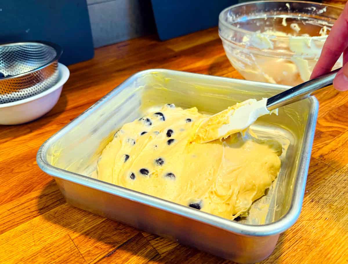 Pale yellow butter studded with blueberries being spread in a square metal baking pan with a silicone spatula.