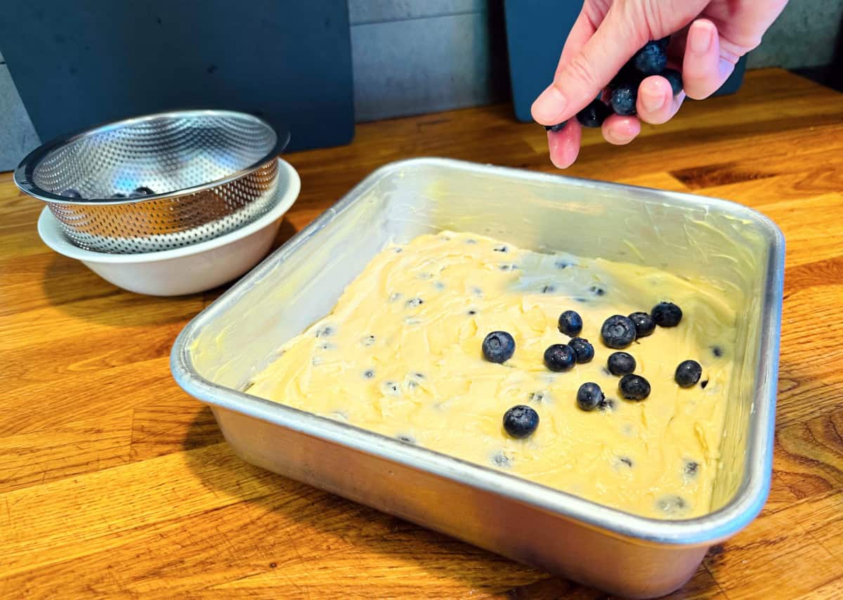 Blueberries being sprinkled over pale yellow batter in a square metal baking pan.