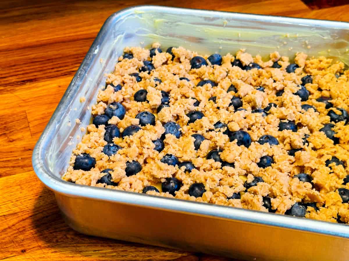 Streusel covered blueberries and batter in a square metal pan sitting on a wooden counter.