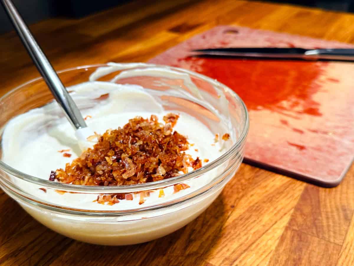 Chopped fried shallots sitting on top of a white creamy mixture in a glass bowl next to a cutting board and chef's knife.