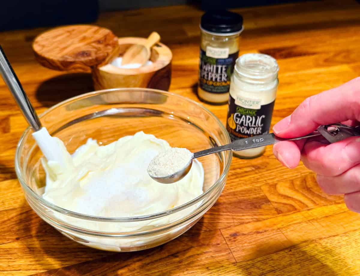 White powder in a steel measuring spoon being held over a small bowl of sour cream and mayonnaise next to a container of salt and jars of white pepper and garlic powder.