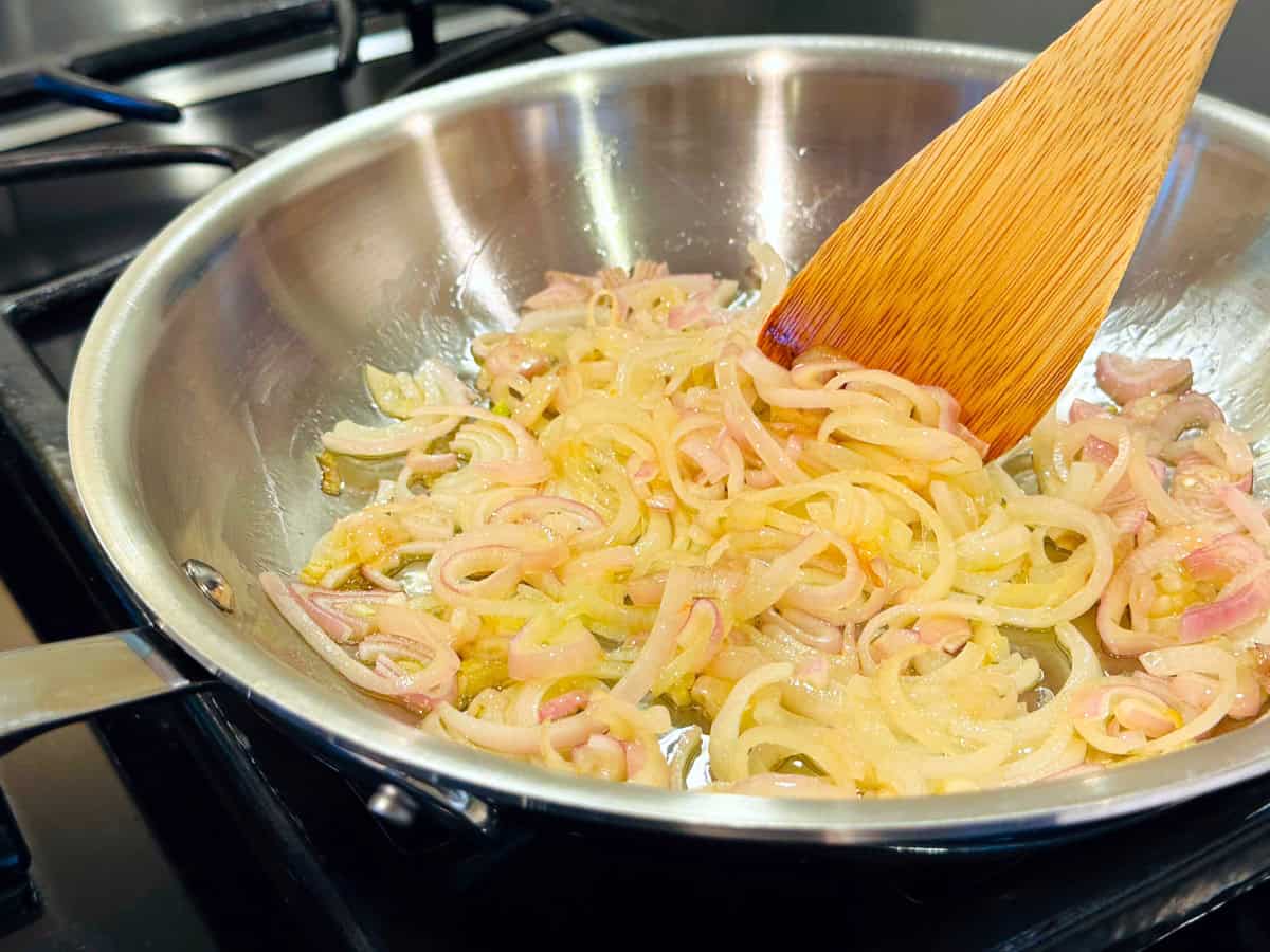 Chopped shallots being stirred with a wooden spatula in a medium steel skillet.