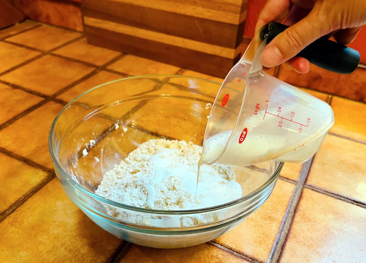 Buttermilk being poured from a plastic measuring cup into a glass bowl containing a sandy mixture of flour and butter.