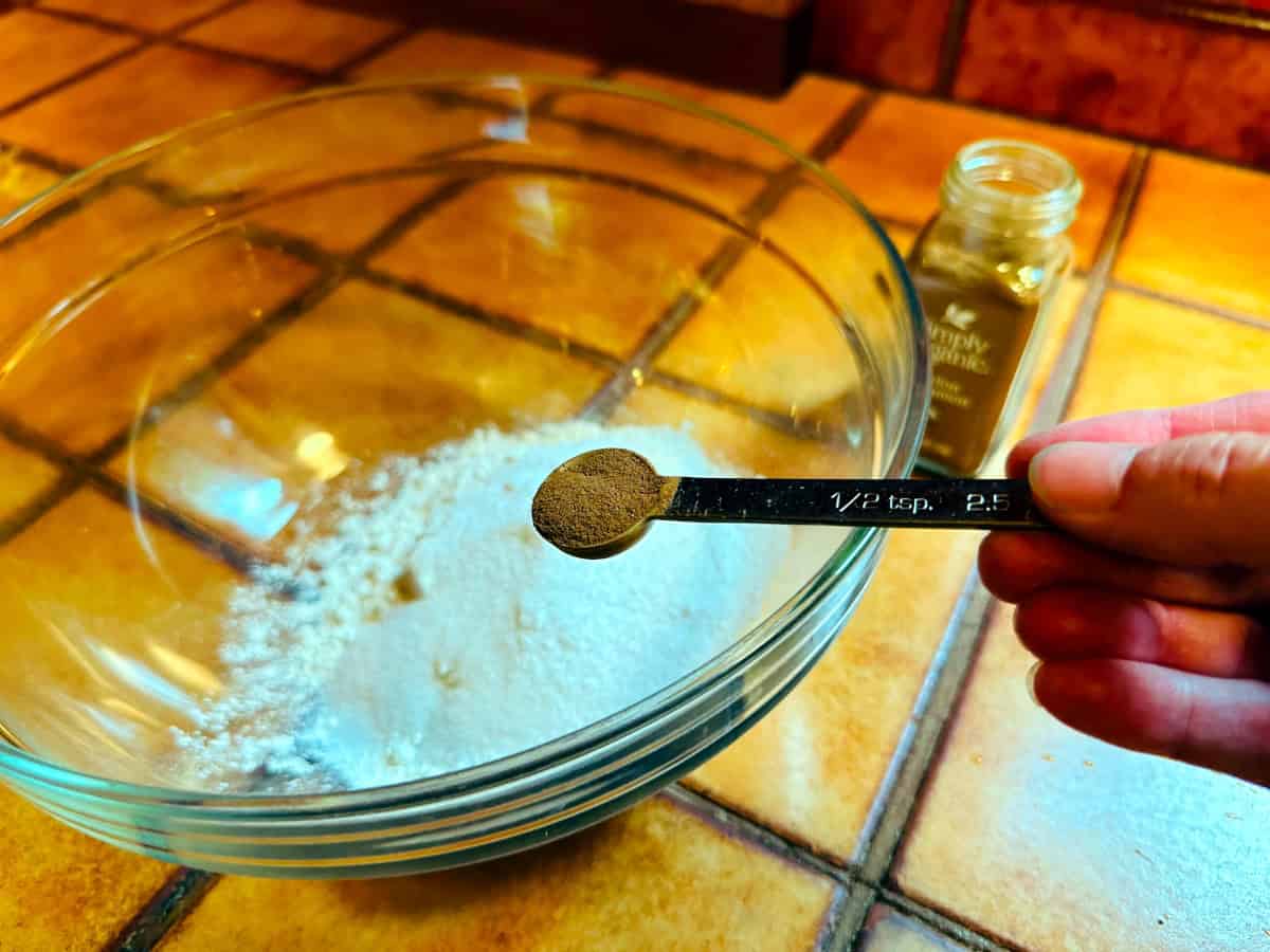 Cinnamon in a steel measuring spoon being held over a glass bowl with flour and sugar.