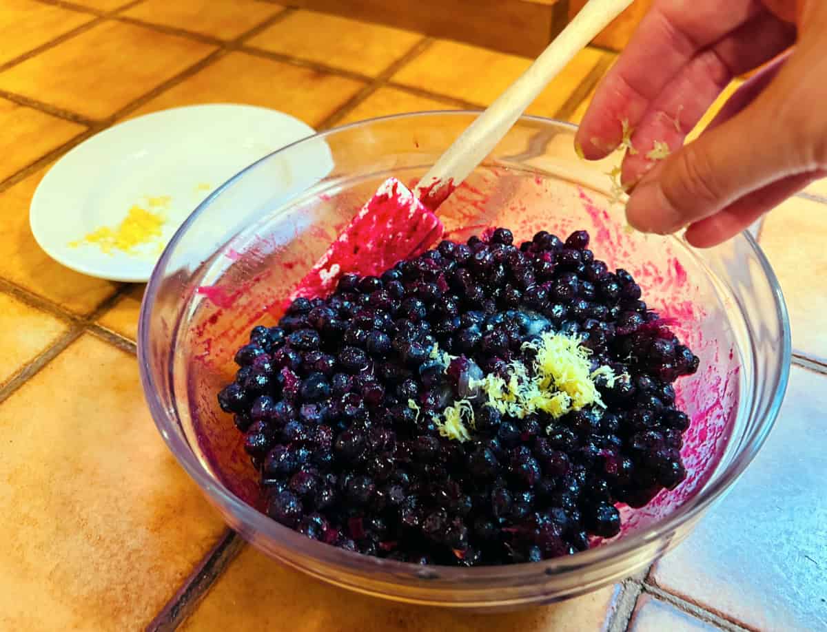 Lemon zest being sprinkled into a glass bowl of blueberries.