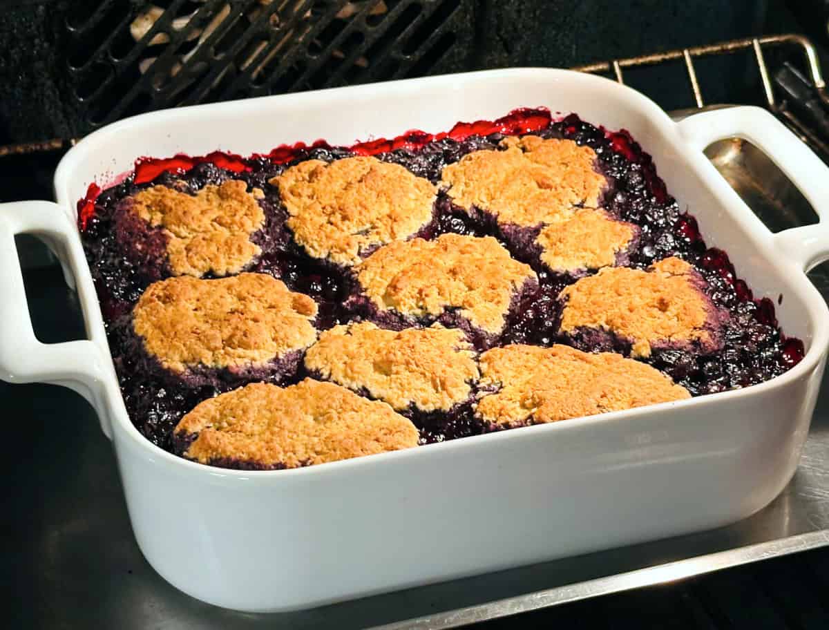 Blueberry cobbler cooking in a square white baking dish in the oven.