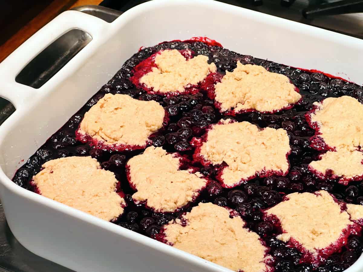 Biscuit dough arranged on top of cooked blueberries in a square white baking dish.