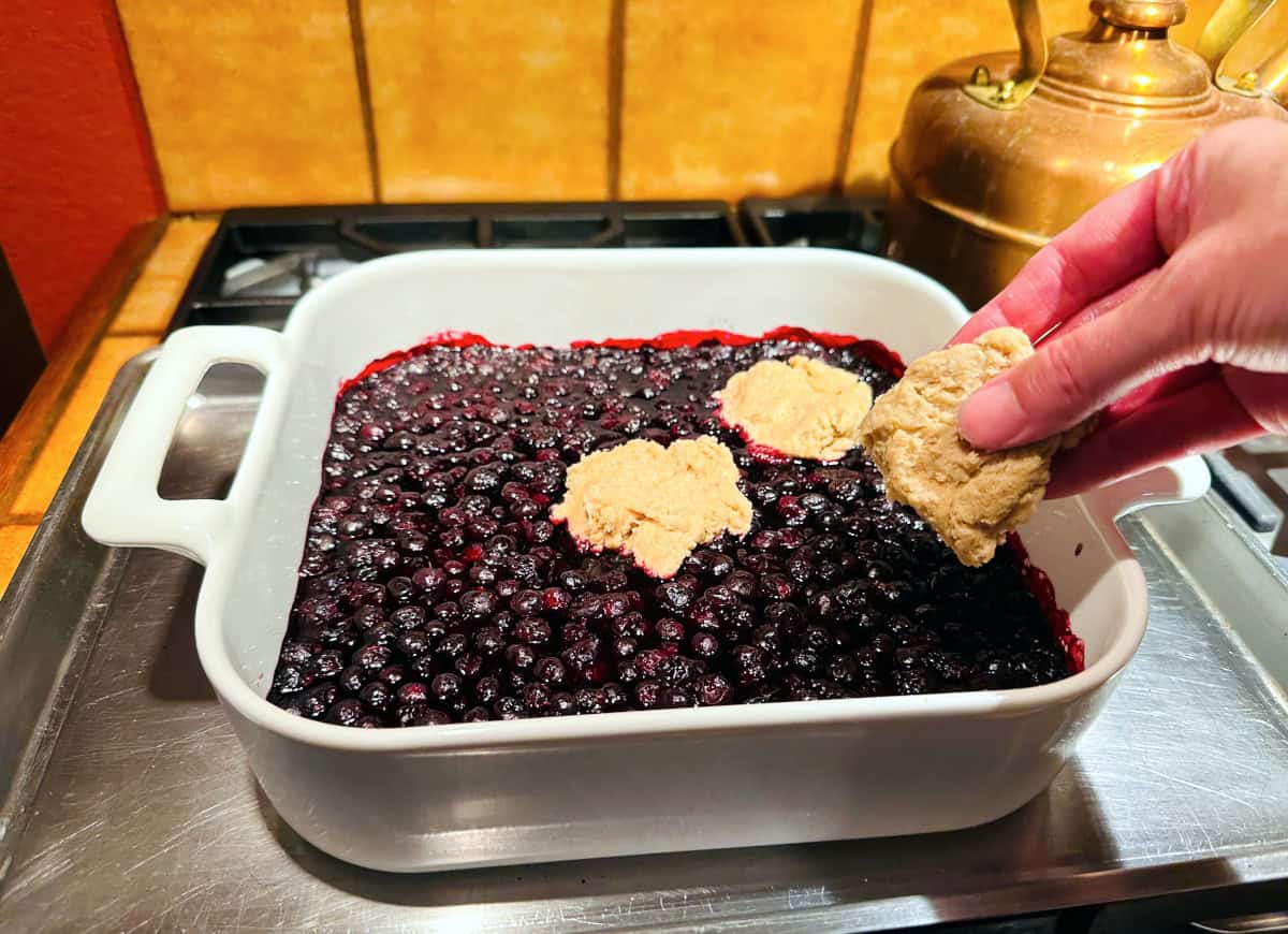 Rough rounds of pale tan biscuit dough being placed on top of cooked berries in a square white baking dish.