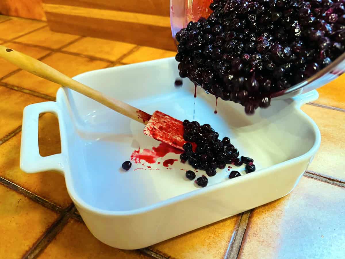 Blueberry mixture being poured from a glass bowl into a square white baking dish with a silicone spatula sitting inside.