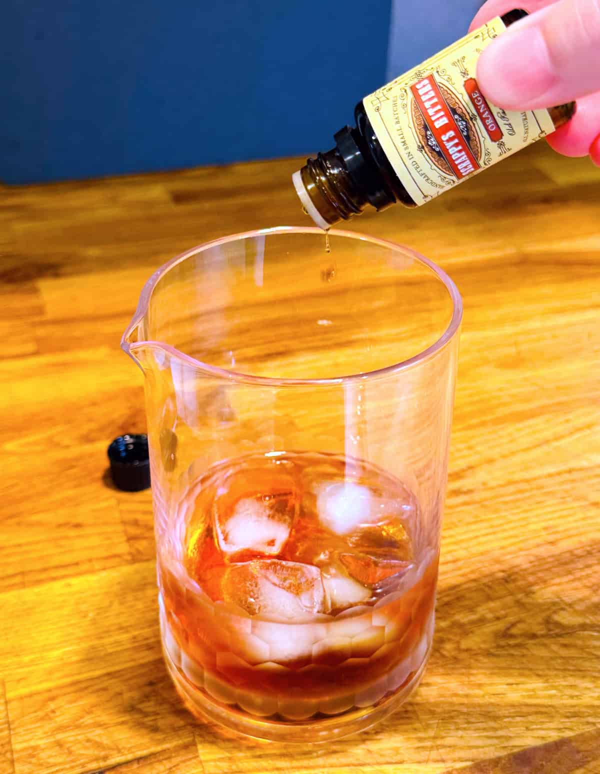 Drop of liquid falling from a small bottle of orange bitters into a mixing glass containing amber liquid and ice.