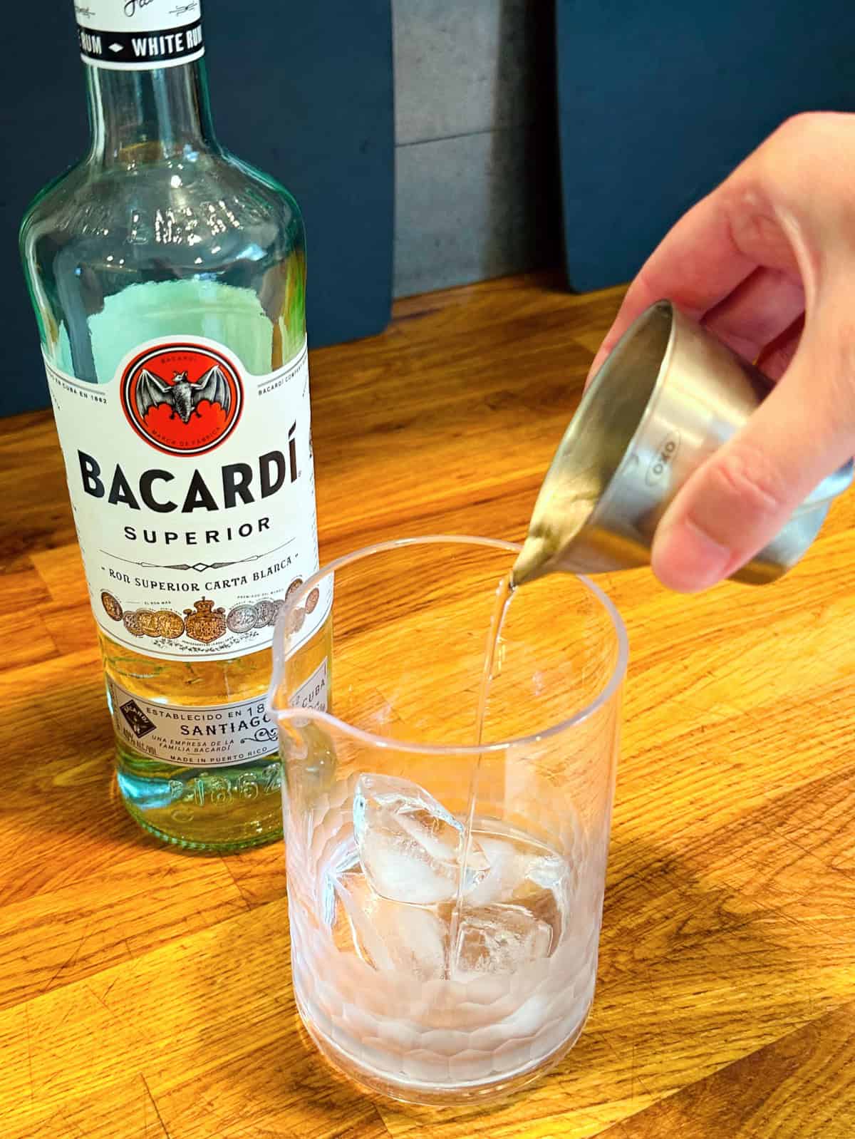 Clear liquid being poured from a steel measuring jigger into a mixing glass with ice next to a bottle of Bacardi rum.