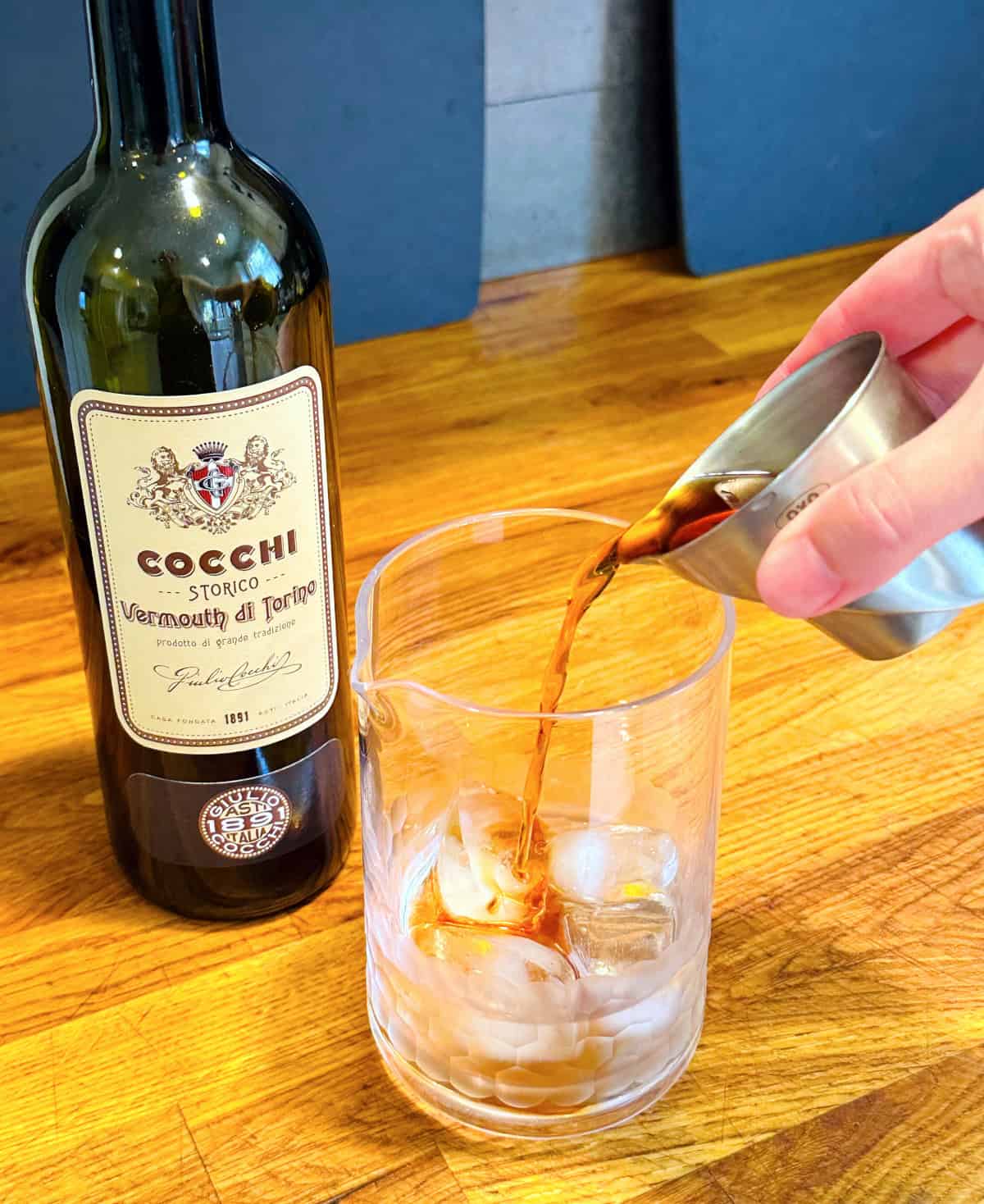 Brown liquid being poured from a steel measuring jigger into a mixing glass with ice next to a bottle of cocchi vermouth di Torino.