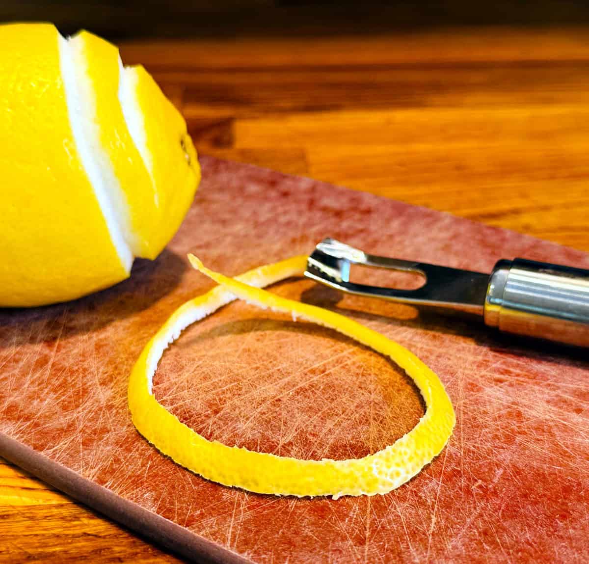 Strip of lemon peel on a cutting board between a steel citrus channel knife and a lemon.