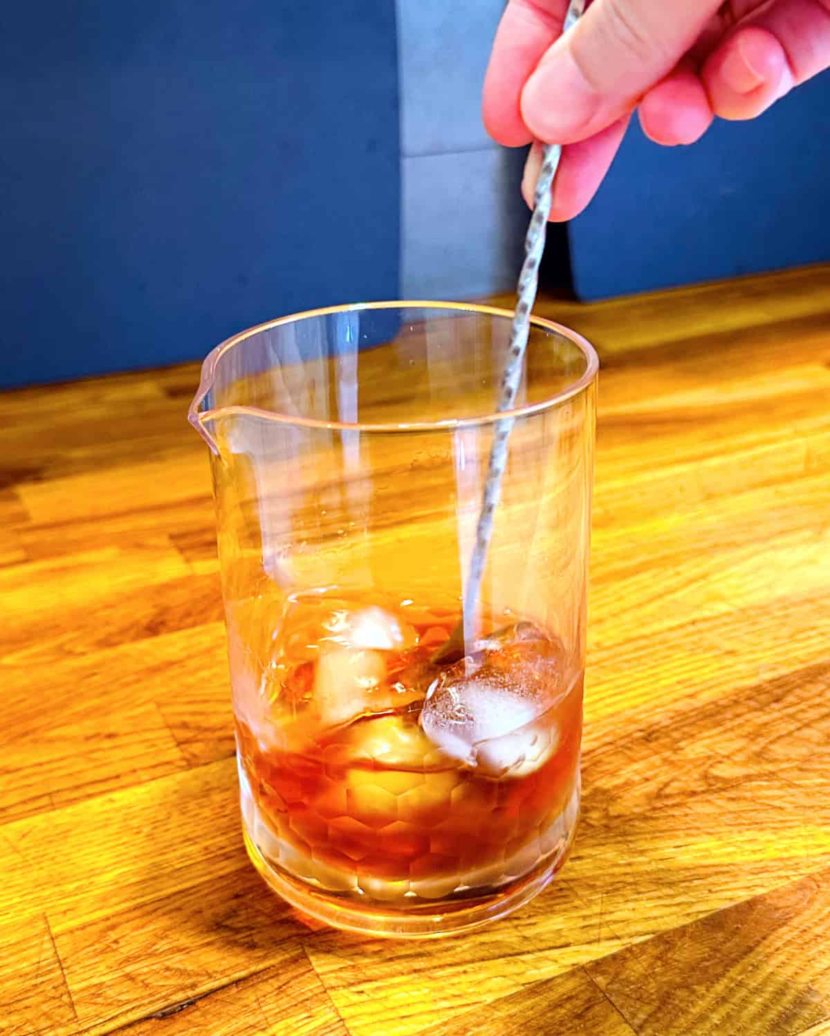 Amber colored liquid and ice being stirred with a metal bar spoon in a mixing glass.