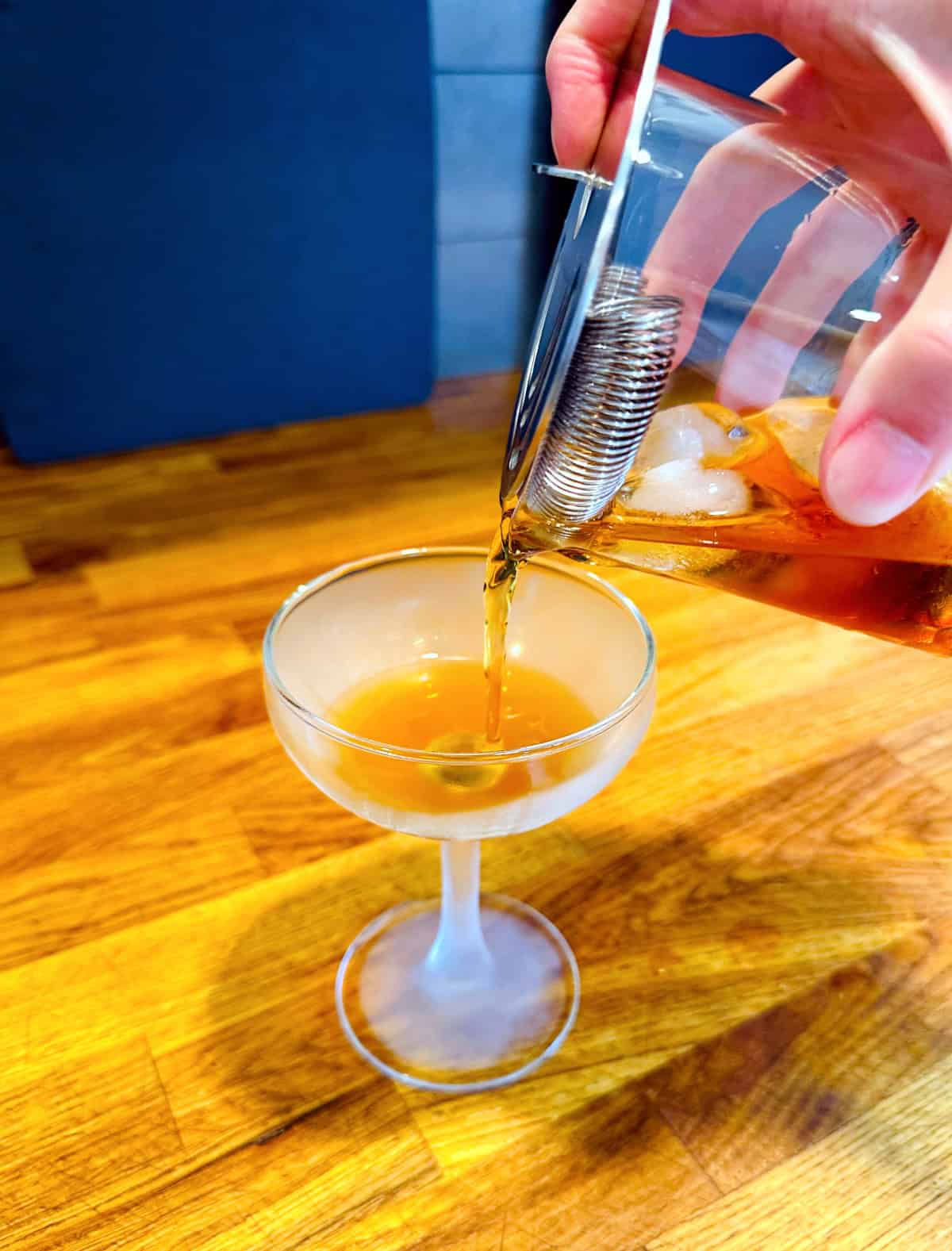 Amber colored liquid being strained from a mixing glass into a chilled coupe glass.