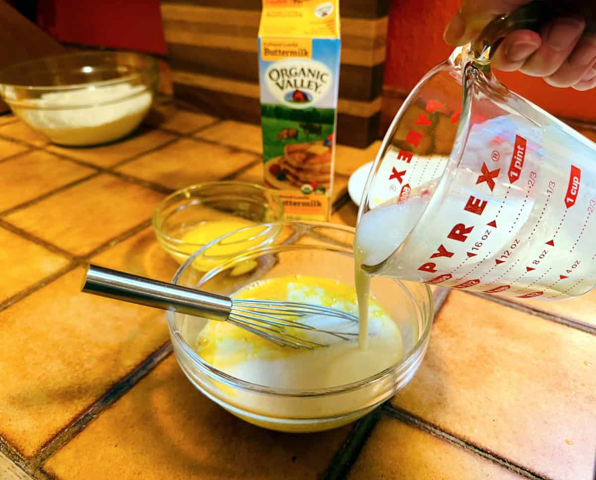 Buttermilk being poured from a glass measuring cup into yellow liquid in a small glass bowl with a metal whisk sitting inside.