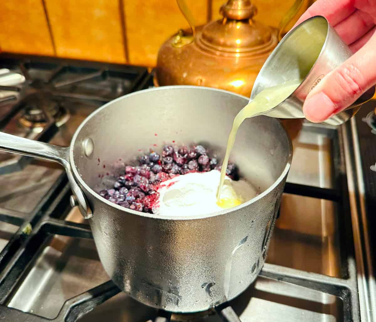 Lemon juice being poured from a steel measuring jigger into a small steel saucepan containing blueberries sugar, and cornstarch.