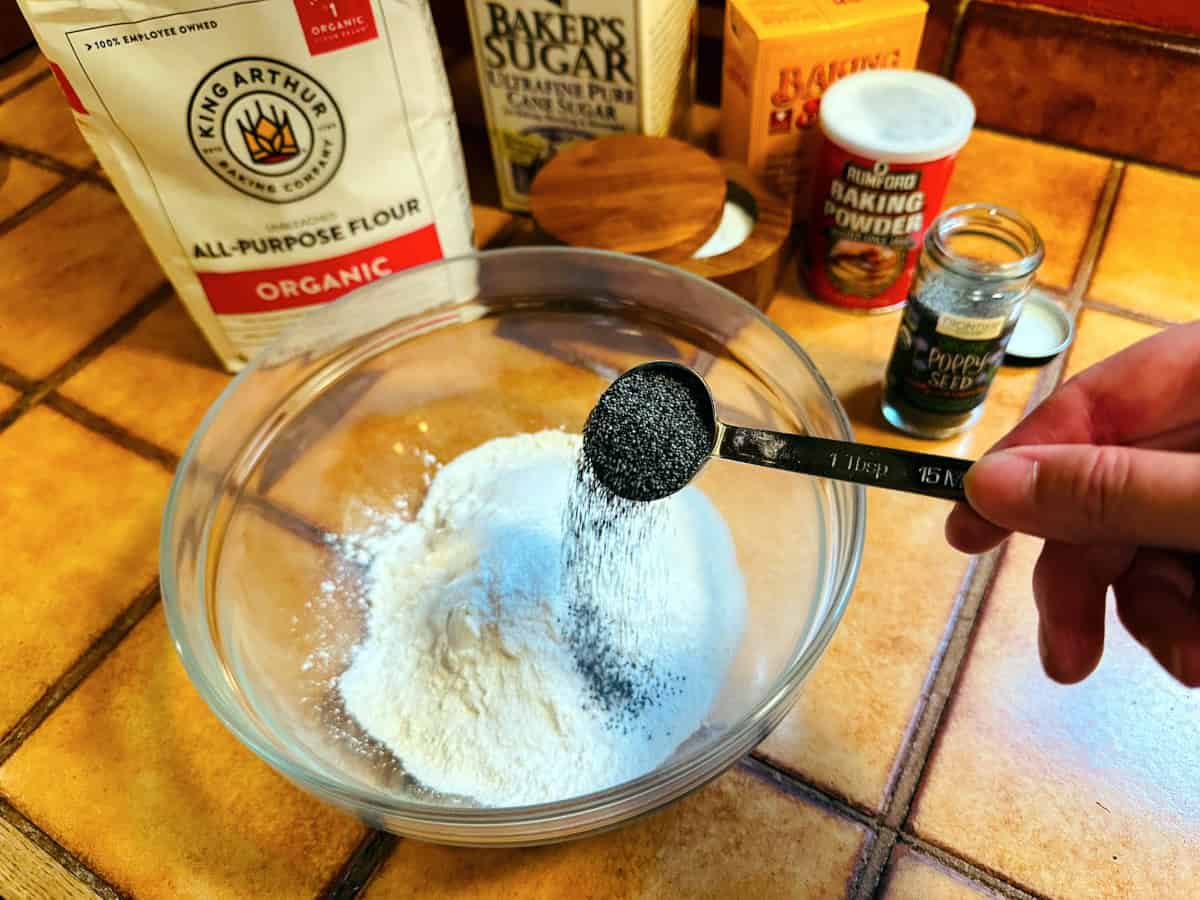 Poppy seeds being tipped from a steel measuring spoon into a glass bowl containing flour.