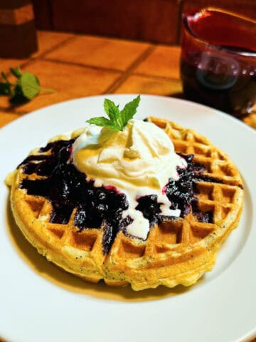 Lemon poppy seed waffle topped with blueberry sauce, whipped cream, and a mint leaf on a white plate.