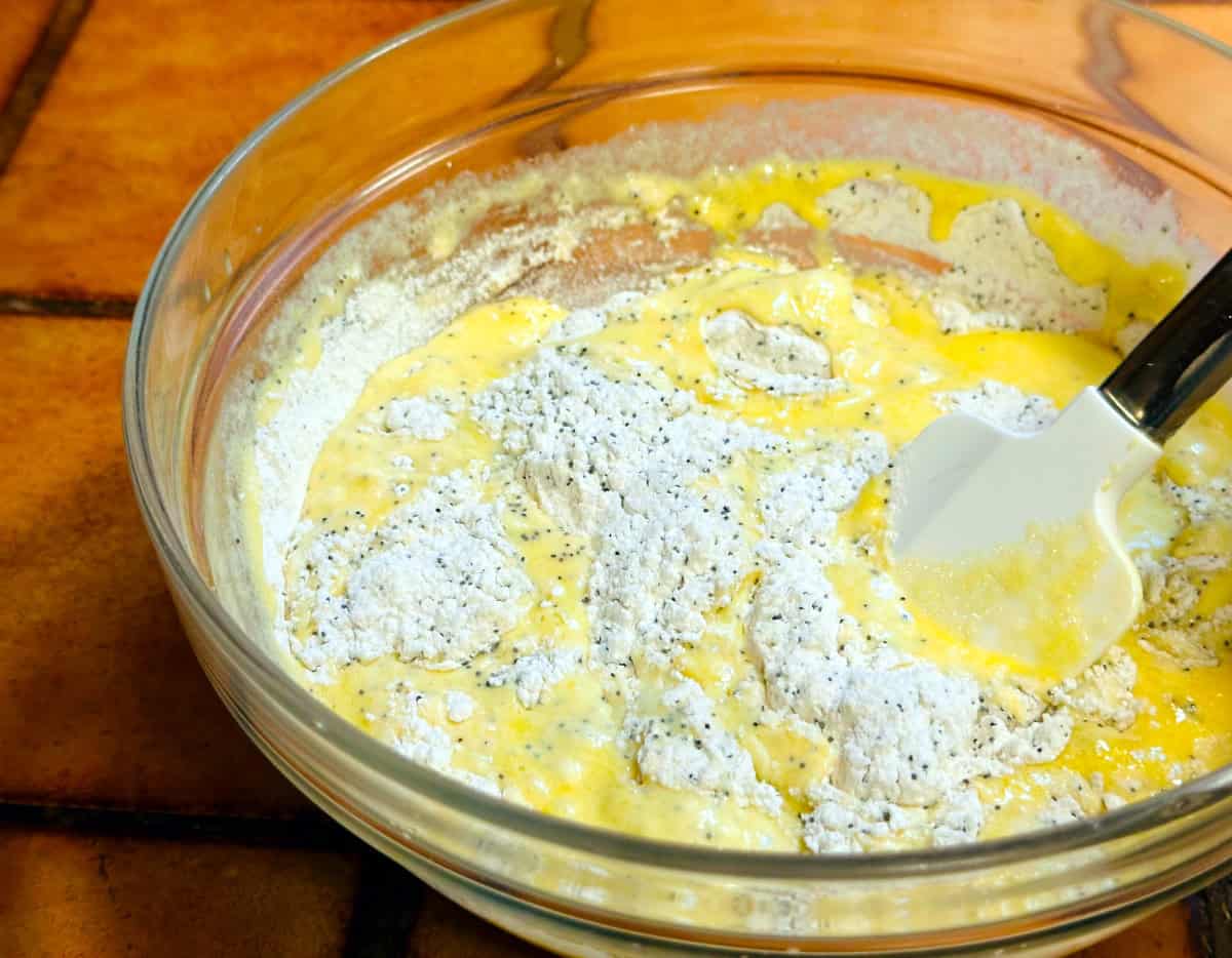 Mixture of yellow liquid and poppy seed studded flour being stirred together in a glass bowl with a white silicone spatula.