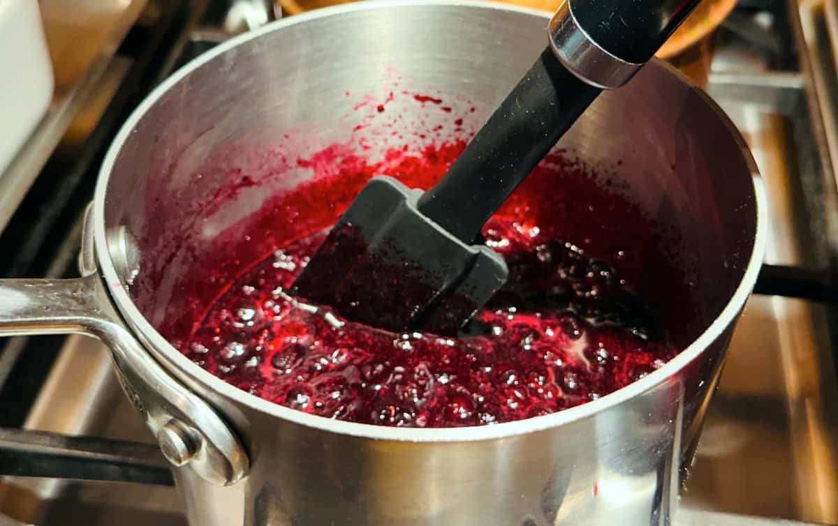 Blueberry sauce simmering on the stove in a small steel saucepan while being stirred with a black silicone spatula.