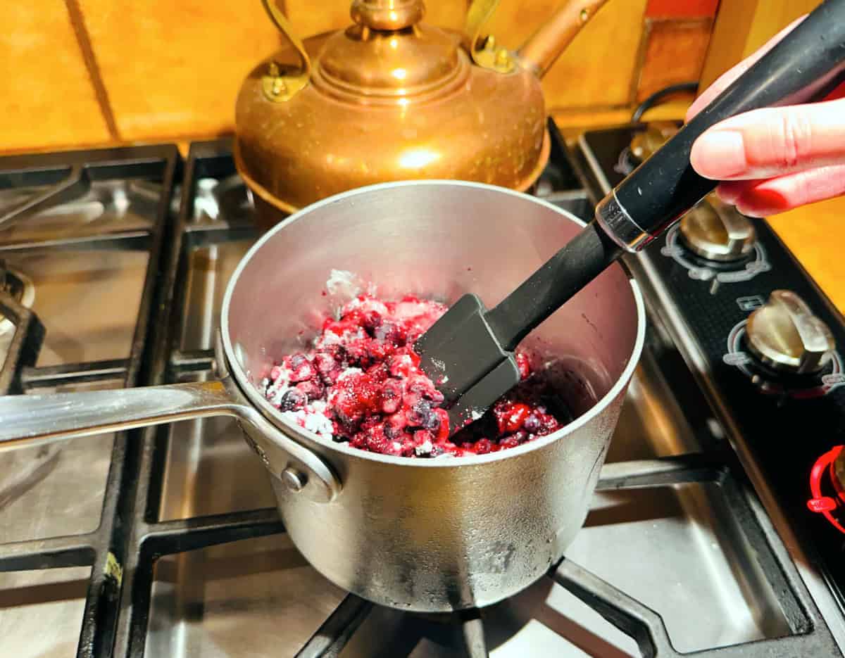 Blueberries, sugar, and cornstarch being stirred together with a black silicone spatula in a small steel saucepan.