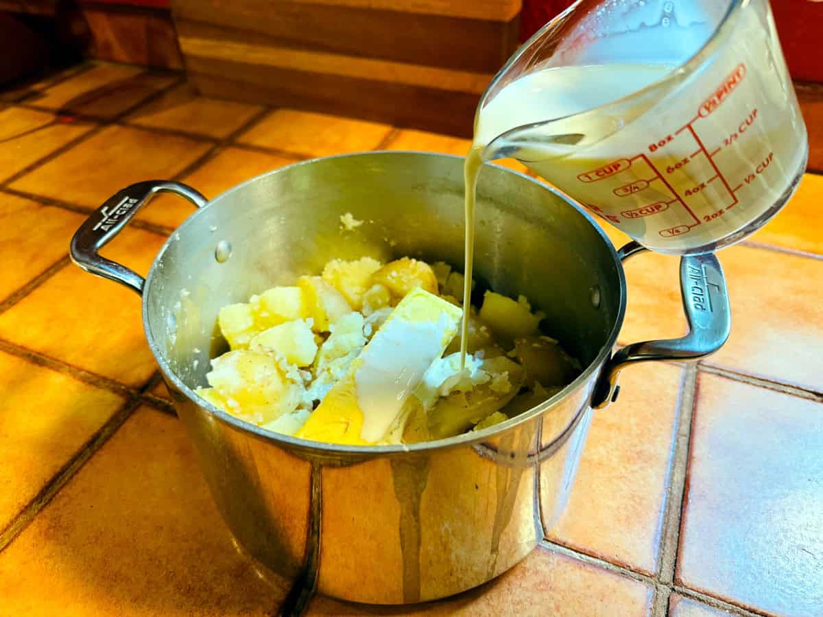 Cream being poured from a glass measuring cup into cooked potatoes and butter in a steel pot.