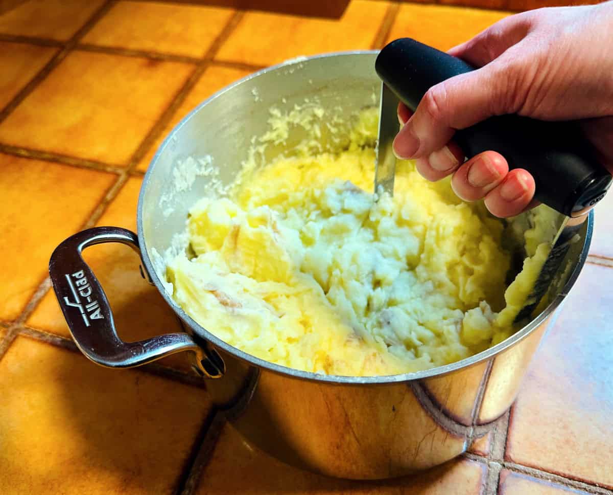 Potatoes being crushed with a hand masher in a steel pot.