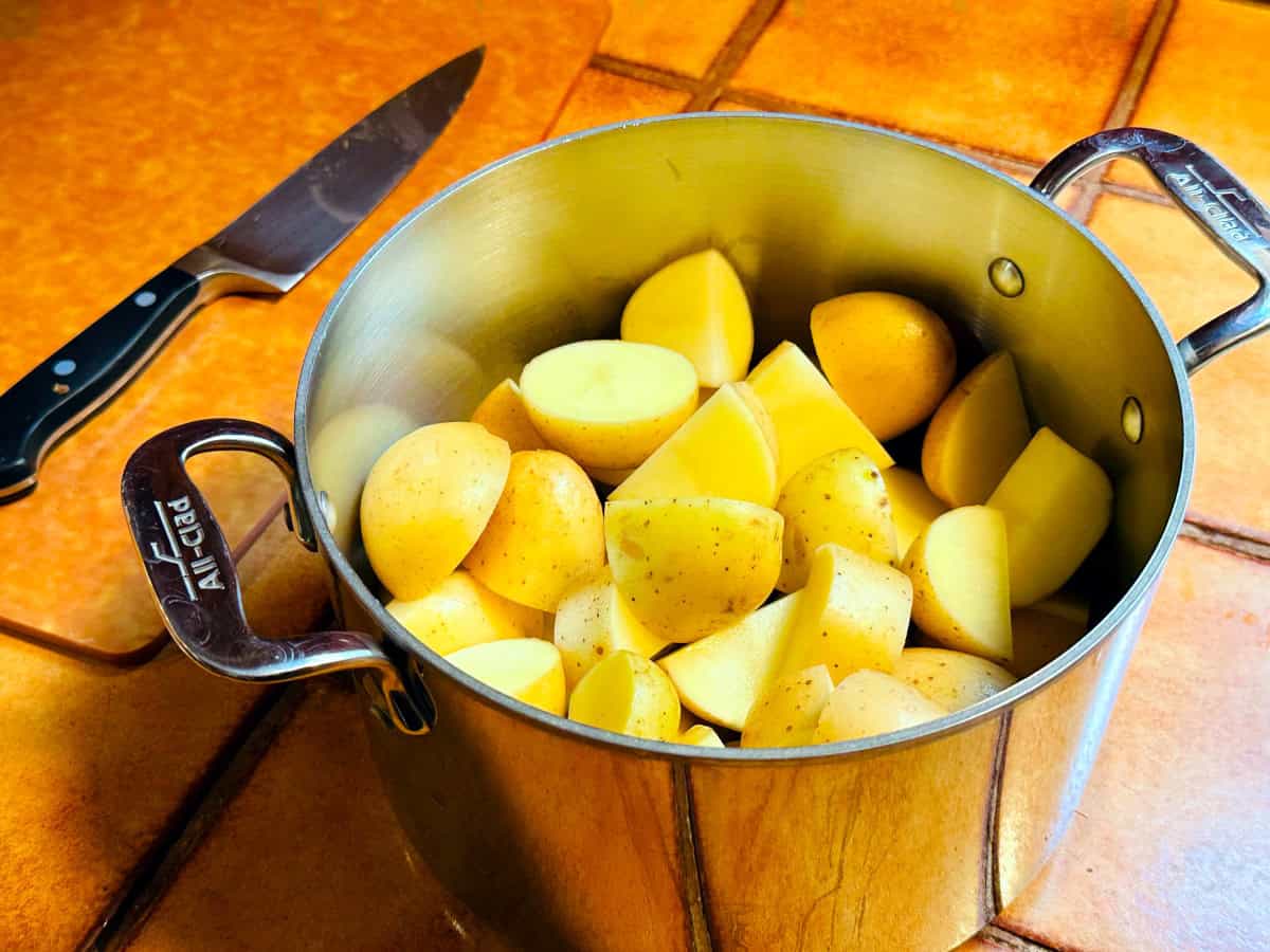 Raw potato chunks in a steel pot next to a chef's knife sitting on a cutting board.