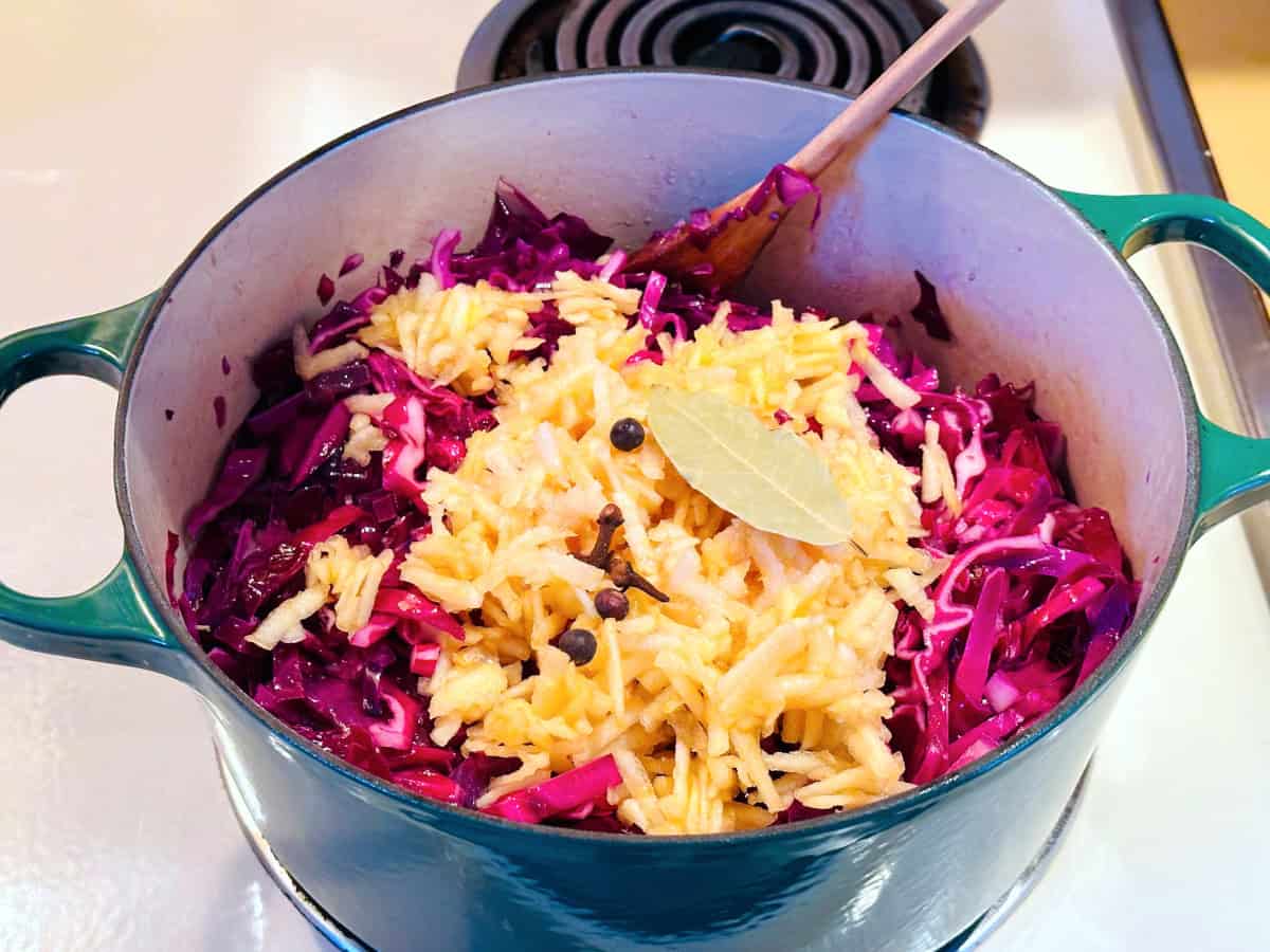 Chopped red cabbage in a dark green dutch oven with grated apple, juniper berries, cloves, and a bay leaf sitting on top.