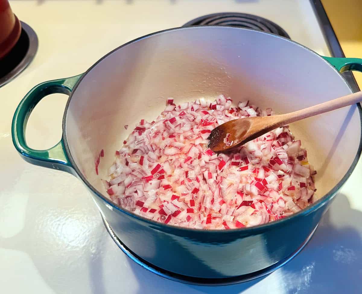 Chopped red onion being stirred with a wooden spoon in a dark green dutch oven.