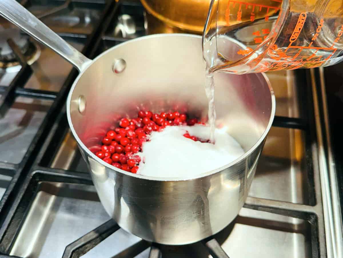 Water being poured from a glass measuring cup over sugar and red berries in a small steel saucepan on the stove.