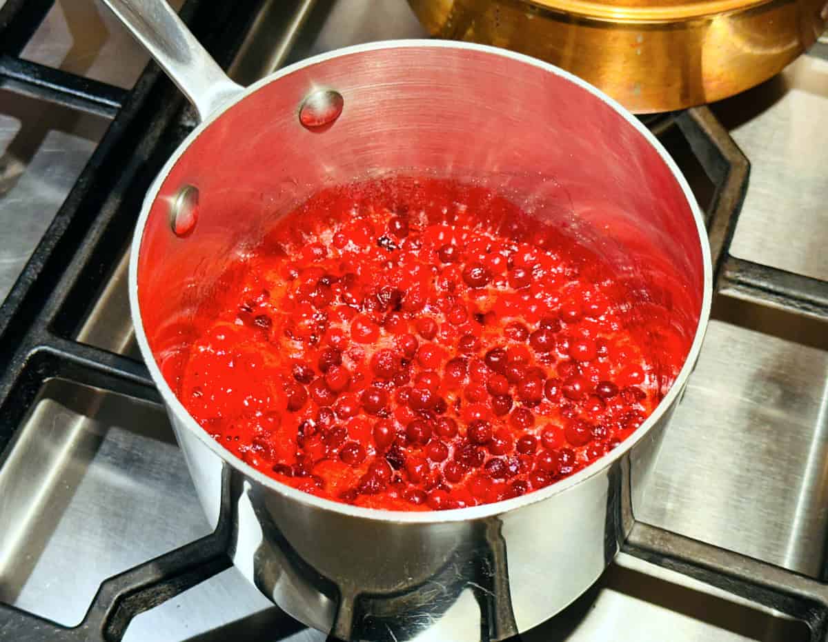 Bright red lingonberries boiling in a small steel saucepan on the stove.