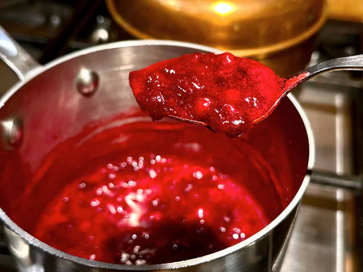 Dark red berry sauce in a spoon being held over a small steel saucepan full of berry sauce.