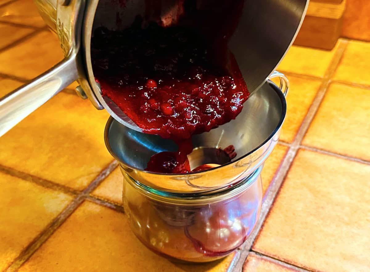 Dark red berry sauce being poured from a small steel saucepan through a metal canning funnel sitting on a glass jar.