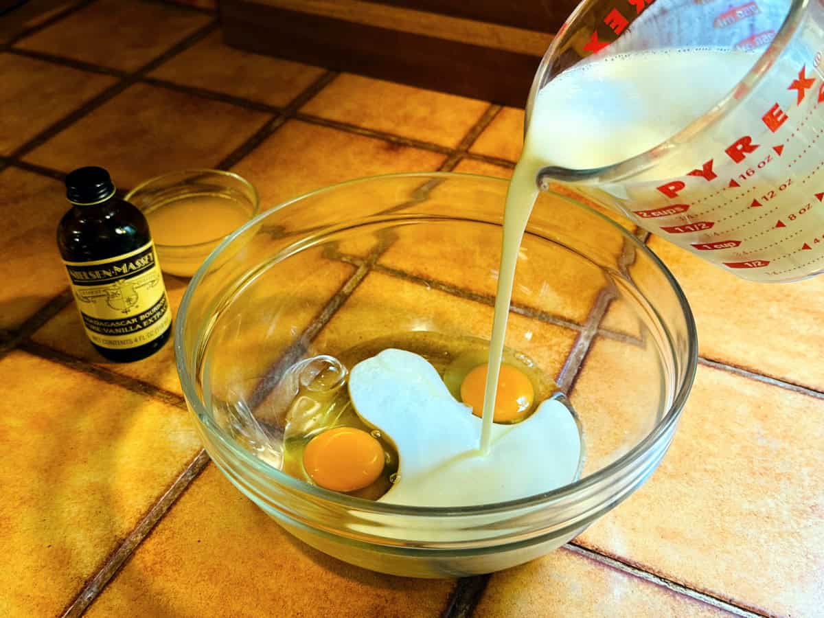 Buttermilk being poured from a glass measuring cup into a glass bowl containing two eggs next to a bottle of vanilla and a small glass bowl of melted butter.