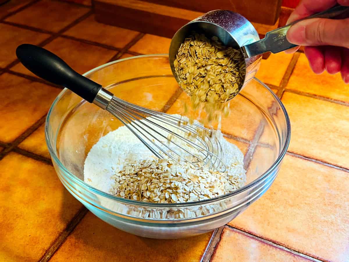 Oats being poured from a steel measuring cup into a glass bowl containing flour and a whisk.