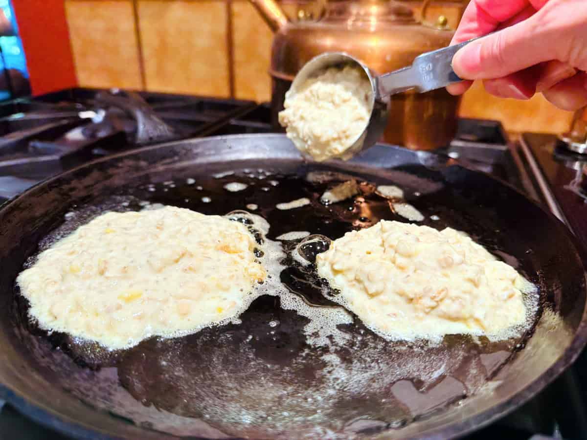 Pancake batter being tipped out of a small steel measuring cup onto a cast iron griddle next to two other dollops of batter.