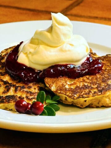 Oatmeal pancakes topped with lingonberry sauce and whipped cream on a white plate.