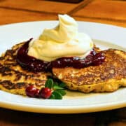 Oatmeal pancakes topped with lingonberry sauce and whipped cream on a white plate.