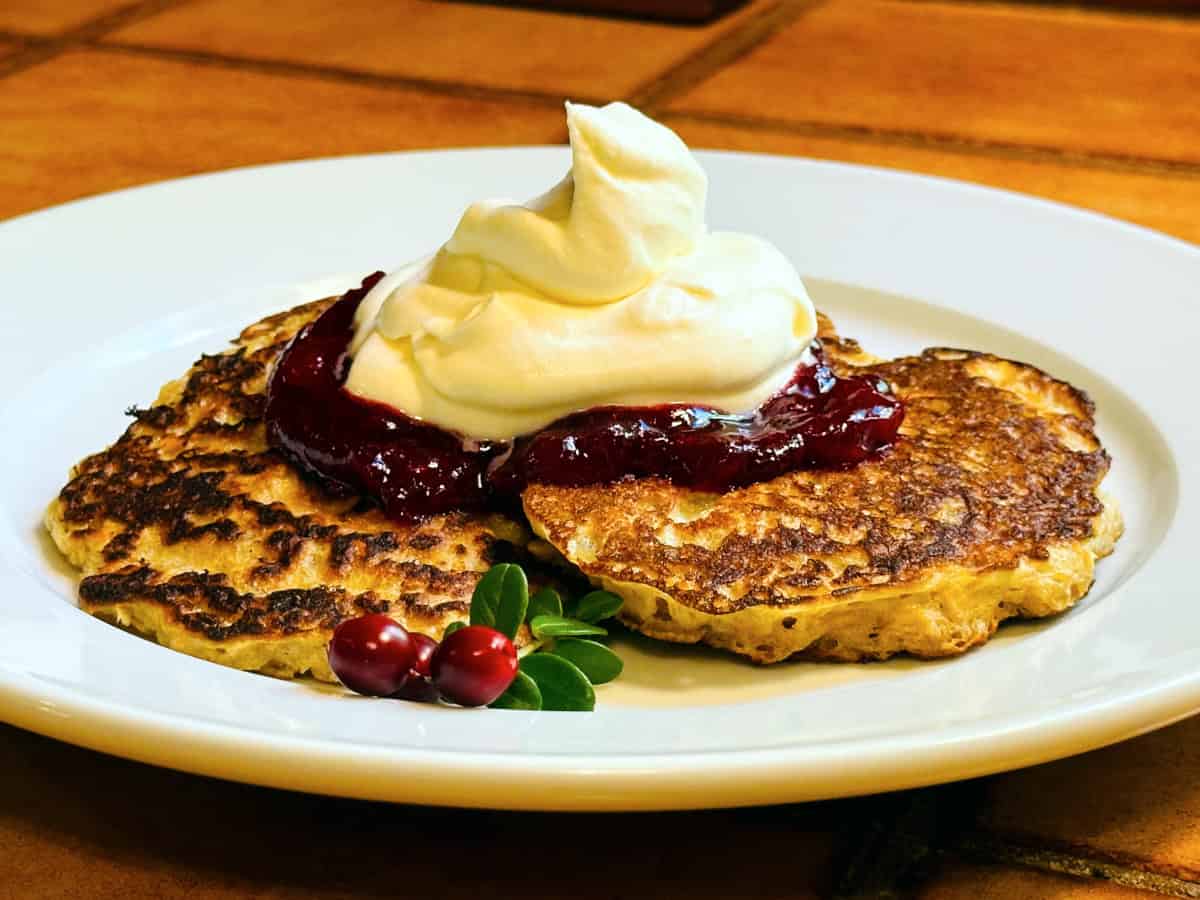 Oatmeal pancakes topped with lingonberry sauce and whipped cream on a white plate.