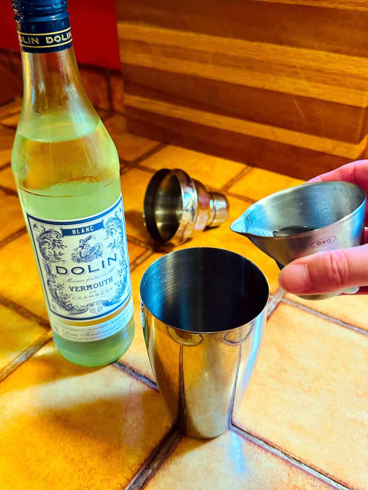 Pale yellow liquid in a steel measuring jigger being held over a cocktail shaker next to a bottle of blanc vermouth.