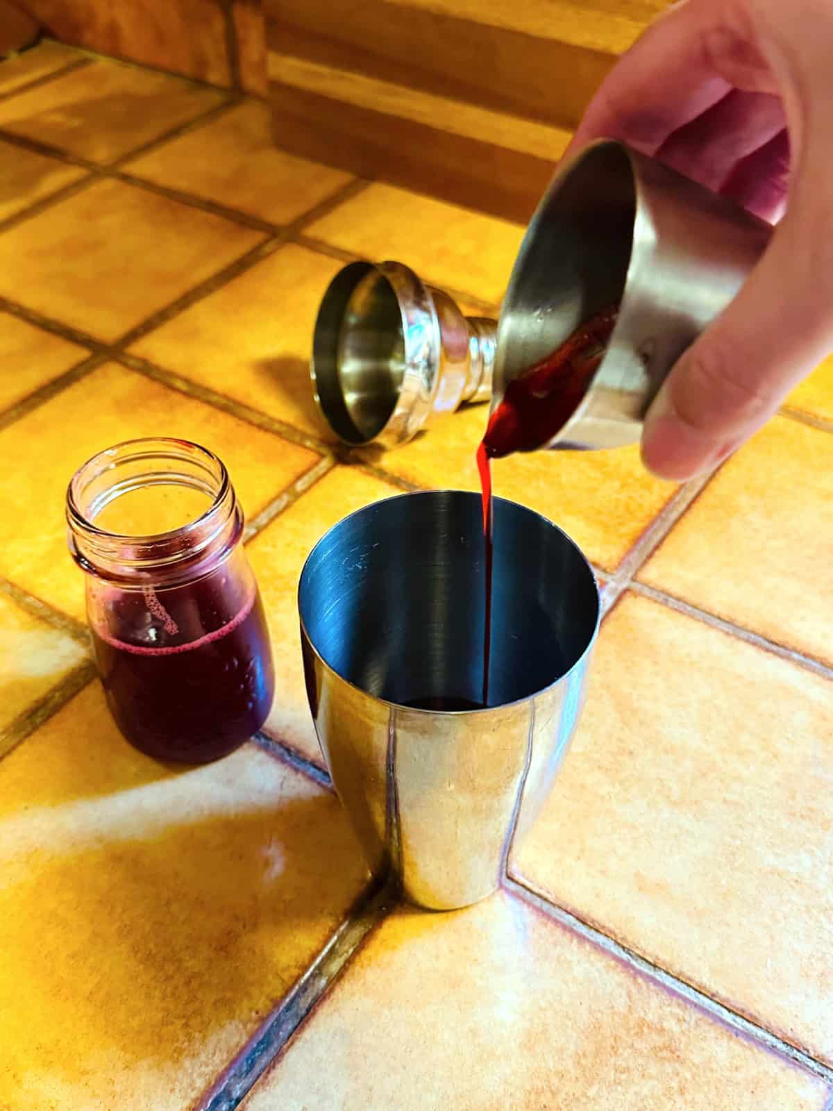 Red liquid being tipped from a steel measuring jigger into a cocktail shaker next to a jar of dark red liquid.