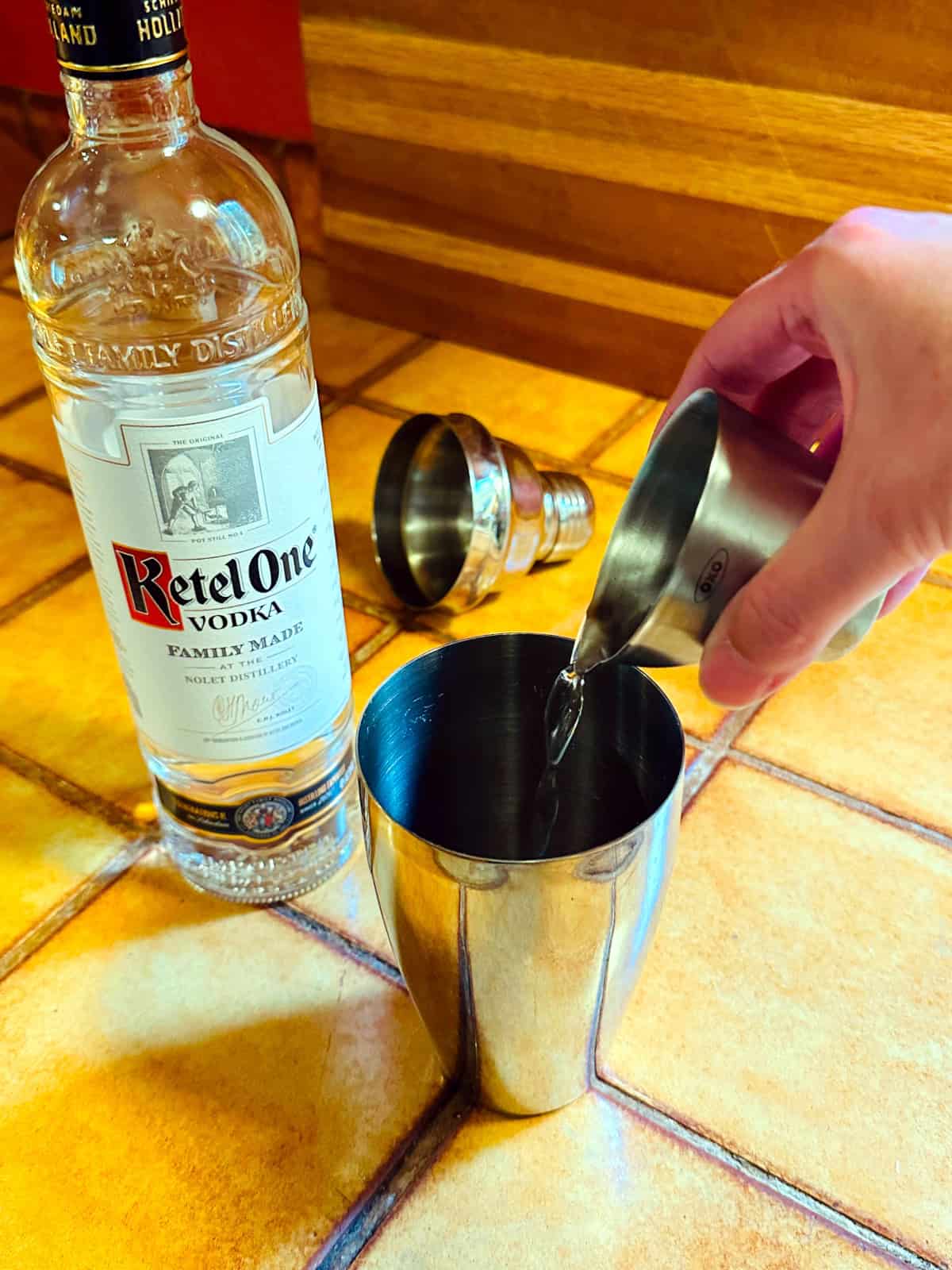 Clear liquid being tipped from a steel measuring jigger into a cocktail shaker next to a bottle of vodka.