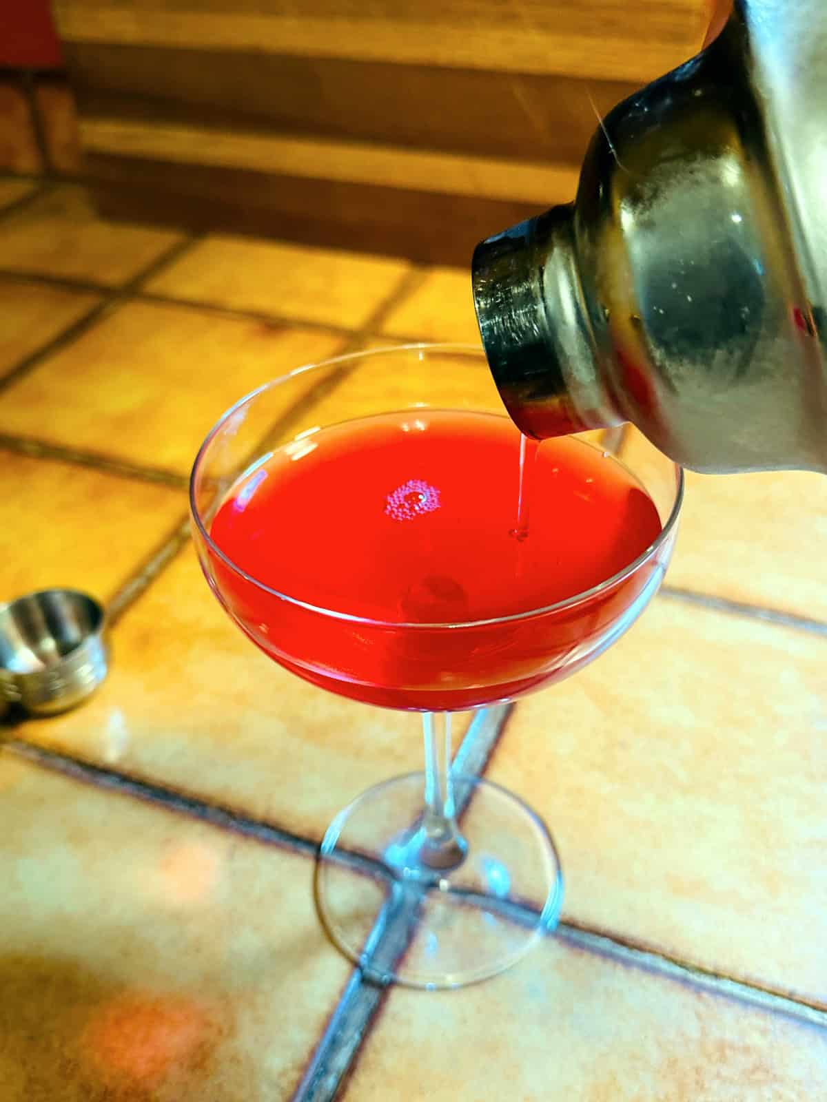 Red liquid being strained from a cocktail shaker into a coupe glass.