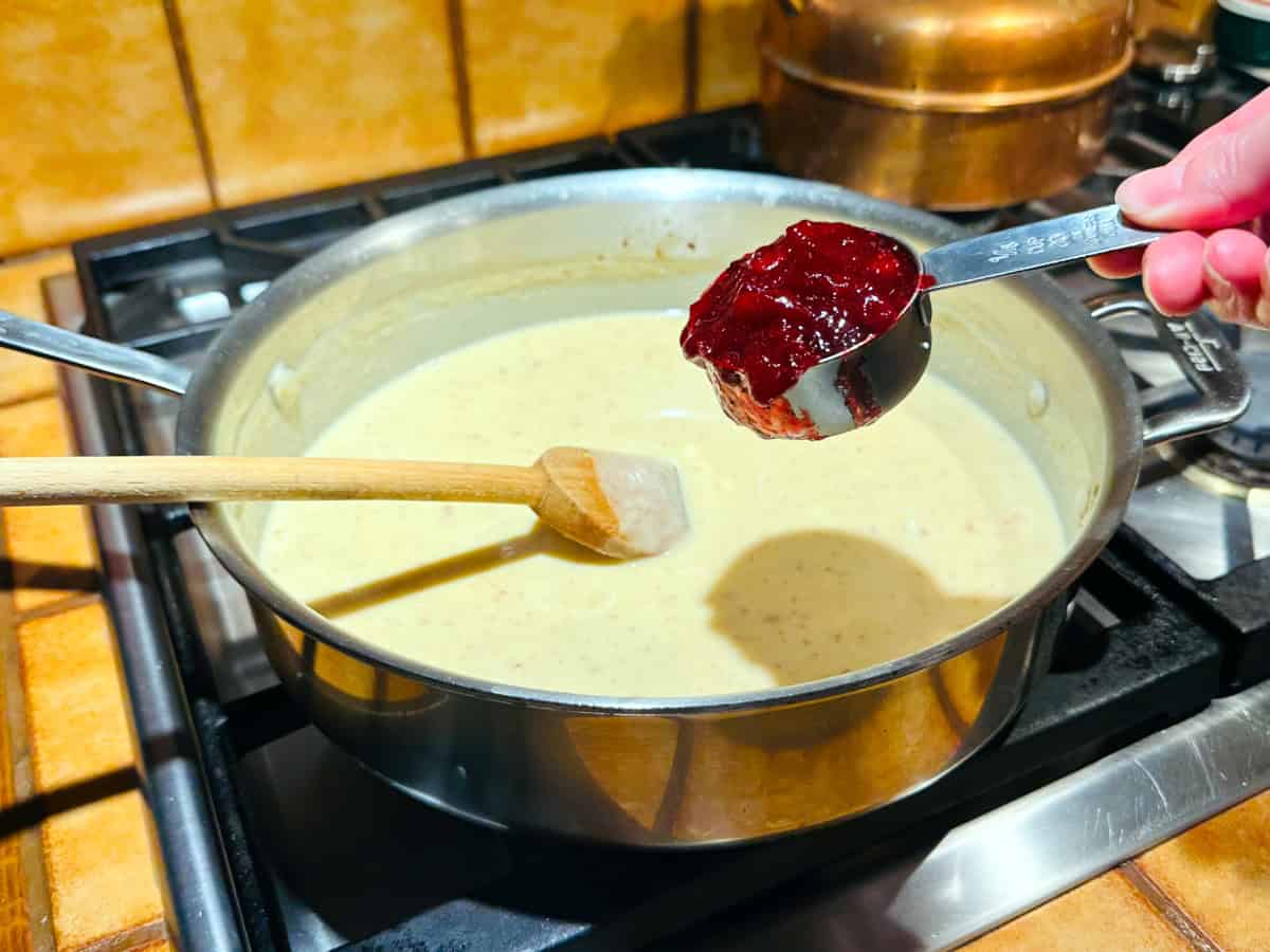 Lingonberry sauce in a steel measuring cup being tipped into cream colored sauce in a deep sided steel saute pan.