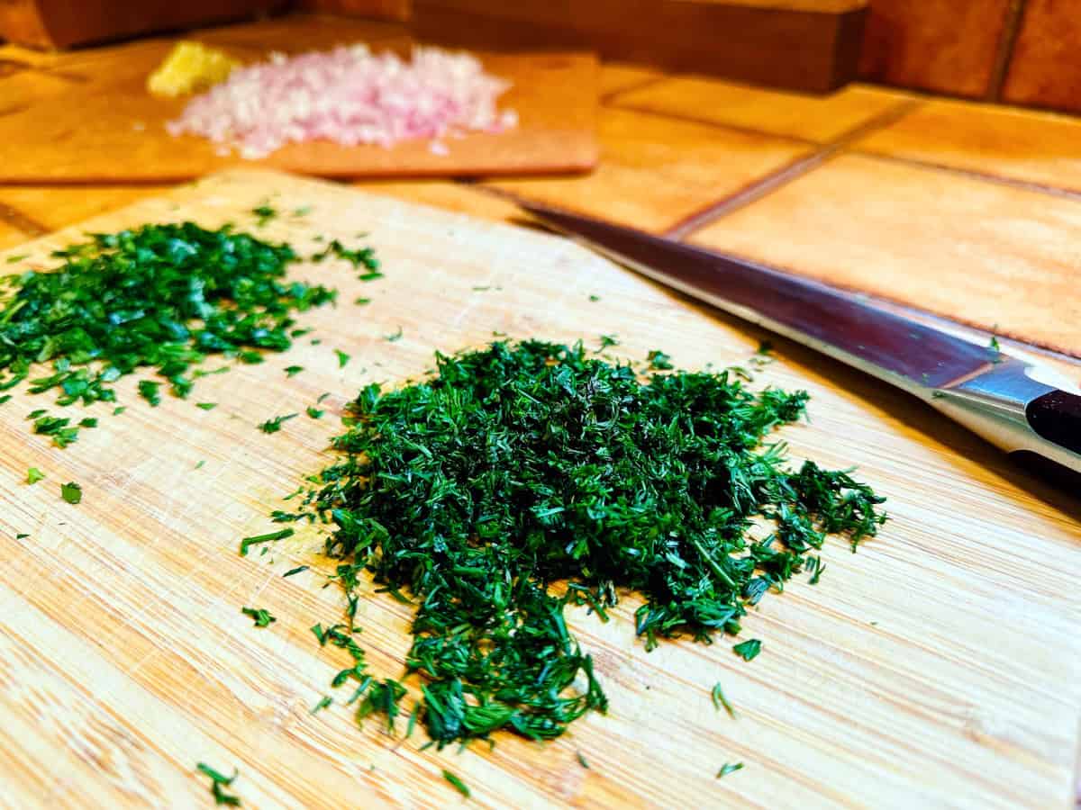 Mound of chopped dill on a cutting board between a mound of chopped parsley and a chef's knife.