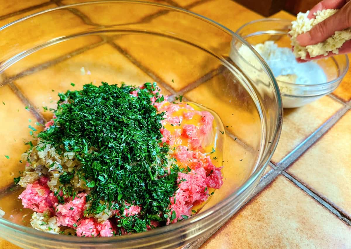 Raw ground meat, shallot mixture, egg, and chopped fresh herbs in a large glass bowl next to a small glass bowl of breadcrumbs soaking in milk.