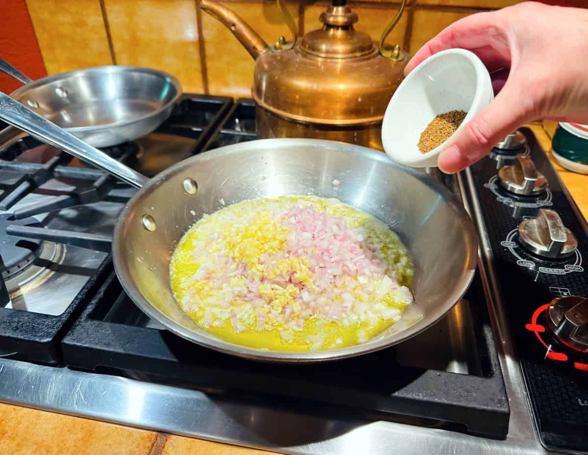 Brown powder being tipped from a small white bowl into a steel frying pan containing chopped shallots and garlic.